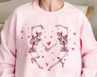 May include: Pink sweatshirt with a graphic of two skeletons dancing and holding hands. The skeletons are surrounded by pink hearts and stars.