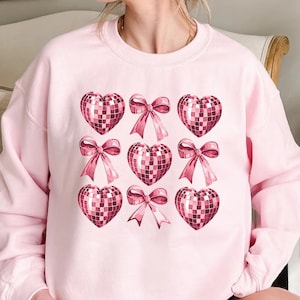 May include: Light pink sweatshirt featuring a pattern of pink disco ball hearts and bows. The design is arranged in a grid pattern across the front of the garment. The sweatshirt is made of a soft material and has a crew neck.