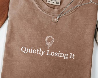 May include: A brown Comfort Colors t-shirt with the text "Quietly Losing It" in white. A silver necklace with a small pendant rests on the shirt. The shirt is size small.