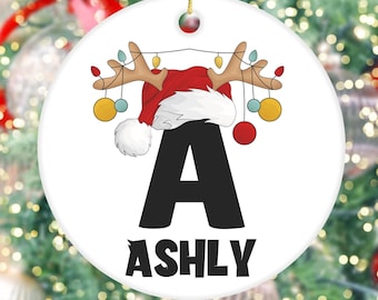 May include: White ceramic Christmas bauble featuring a red Santa hat and reindeer antlers with fairy lights. The letter A and the name ASHLY are printed on the bauble.