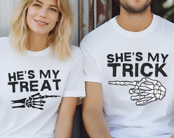 May include: Two white t-shirts with black text and skeleton hand graphics. One shirt reads "HE'S MY TREAT" and the other says "SHE'S MY TRICK". The shirts are likely for a couple, themed for Halloween.