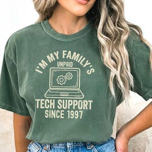 May include: Green t-shirt with the text "I'M MY FAMILY'S UNPAID TECH SUPPORT SINCE 1997" and a laptop graphic. The shirt is a solid green color and has a crew neck.