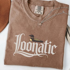 May include: A brown short-sleeved t-shirt with a crew neckline and a vintage-style wash. The front features a graphic of a loon bird above the text "Loonatic" in a stylised white font. A small silver-toned necklace with a circular pendant rests on top.