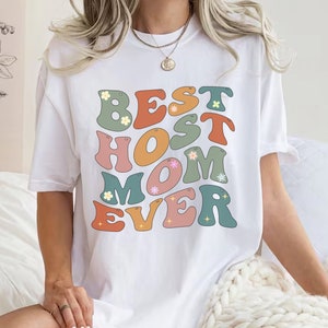 May include: White t-shirt featuring a retro-style graphic that reads "Best Host Mom Ever" in colourful letters with flower and glitter accents.