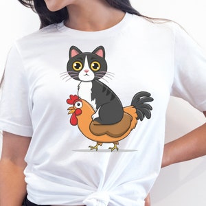 May include: White t-shirt featuring a cartoon illustration of a black and white cat riding a brown and orange chicken.