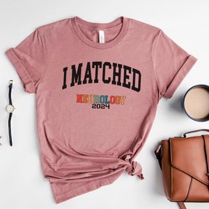 May include: A pink t-shirt with the text "I MATCHED NEUROLOGY 2024" printed on it. The word "NEUROLOGY" is in rainbow colours.