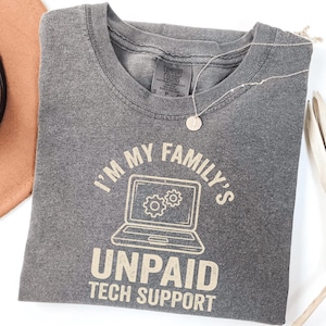May include: A gray Comfort Colors t-shirt with the text "I'M MY FAMILY'S UNPAID TECH SUPPORT" and a laptop graphic. A silver necklace with a small pendant is draped across the shirt.