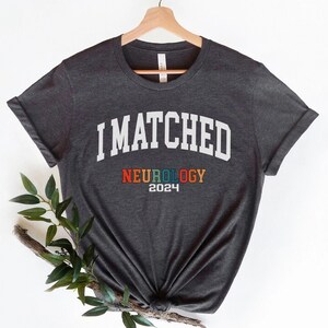 May include: A dark grey t-shirt with the text "I MATCHED" in white, and "NEUROLOGY 2024" in rainbow colours.
