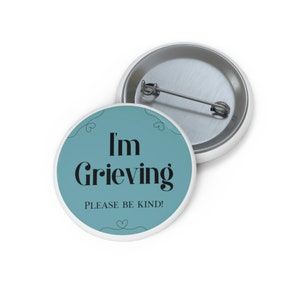 May include: A teal blue button with white trim and the text "I'm Grieving Please Be Kind!" in black. The button has a silver pin on the back.