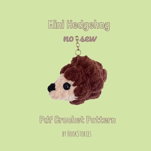 Puede incluir: Un peluche de erizo en miniatura tejido a ganchillo con el lomo texturizado en marrón oscuro y la cara en beige claro, con nariz y ojos negros. El texto dice Mini Hedgehog no sew Pdf Crochet Pattern BY HOOKSTORIES. Incluye una anilla metálica.