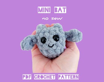 Op de afbeelding: Een gehaakte grijze mini-vleermuis met zwarte ogen en een lachende mond. De vleermuis heeft uitgestrekte vleugels en is gelabeld met "MINI BAT no sew" en "PDF CROCHET PATTERN BY HOOKSTORIES" in witte tekst tegen een paarse achtergrond.