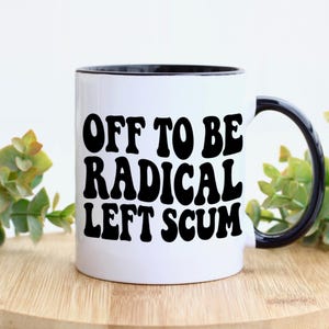 May include: White ceramic mug with a black rim and handle. The mug features the text "OFF TO BE RADICAL LEFT SCUM" in bold, black letters. The mug is sitting on a wooden surface.