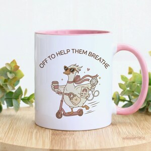 May include: A white ceramic mug with a pink interior and handle, featuring a cartoon goose wearing sunglasses, riding a scooter, and holding a coffee cup and a medical device. The text reads OFF TO HELP THEM BREATHE in brown lettering.