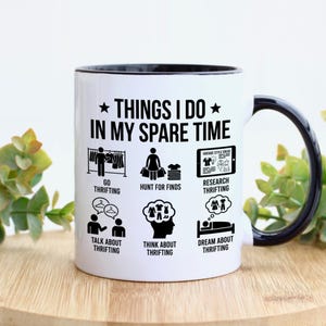 May include: White ceramic mug with a black rim and handle. The mug features the text "THINGS I DO IN MY SPARE TIME" with illustrations of thrifting activities. The mug is on a wooden surface.