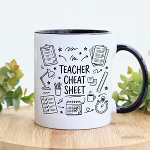 May include: White ceramic mug with a black interior and handle. The mug features a black and white doodle design with the words "Teacher Cheat Sheet" and illustrations of school supplies, including a checklist, lamp, and clock.