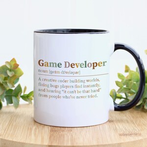 May include: A white ceramic mug with a black handle and interior rim. The front features the text Game Developer, noun, phonetic spelling, and a definition describing a creative coder building worlds, fixing bugs, and hearing common complaints.