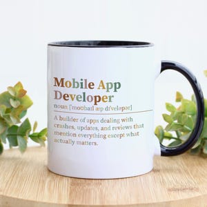 May include: A white ceramic mug with a black interior and handle. The front features the text Mobile App Developer, noun, followed by a phonetic pronunciation and a definition describing a builder of apps dealing with crashes and reviews.