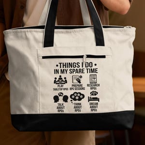 May include: A white tote bag with a black bottom and handles. The bag features the text "THINGS I DO IN MY SPARE TIME" and icons representing tabletop RPG activities like playing, preparing, and researching RPGs.