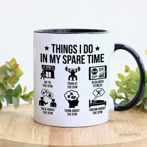May include: White ceramic mug with a black interior and handle. The mug features the text "THINGS I DO IN MY SPARE TIME" and various icons related to fitness, including going to the gym, training, and researching fitness.