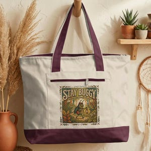 May include: Cream-colored tote bag with a burgundy base, trim, and handles. The front pocket features a colorful illustration of a cartoon bug with the text "STAY BUGGY appreciate small things". The bag is hanging on a wooden hook.