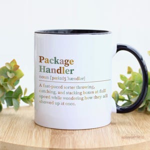 May include: A white ceramic mug with a black interior and handle. The front features the text 'Package Handler' in brown and green, followed by the phonetic spelling and a definition describing a fast-paced sorter throwing and stacking boxes.