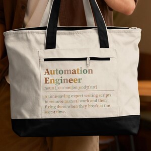 May include: A cream-colored canvas tote bag with black handles and a black base. The front features a zippered pocket printed with the text: Automation Engineer noun [ɔːtəˈmeɪʃən ˌendʒɪˈnɪər] A time-saving expert writing scripts to remove manual work.