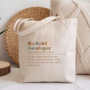 May include: A cream-colored canvas tote bag featuring the printed text: Backend Developer noun [bækˌend dɪˈveləpər] A logic builder handling data, servers, and performance while quietly carrying the entire system nobody sees, in multi-colored lettering.