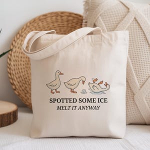 May include: Cream-colored tote bag with a whimsical duck illustration. The design features three ducks and the text "SPOTTED SOME ICE MELT IT ANYWAY" in black. The bag has a simple, casual design.