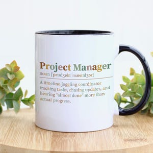 May include: A white ceramic mug with a black handle and interior rim. The mug features the text Project Manager, followed by a phonetic pronunciation and a humorous definition describing a timeline-juggling coordinator tracking tasks.