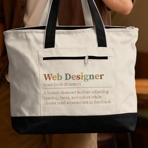 May include: A cream-colored canvas tote bag with black handles and a black base. The front features a zippered pocket and printed text: Web Designer noun [web di'zainer] A layout-obsessed builder adjusting spacing, fonts, and colors while clients send screenshots as feedback.