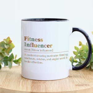 May include: A white ceramic mug with a black handle and interior. The front features the text Fitness Influencer, defined as a content-creating motivator filming workouts, retakes, and angles until it looks effortless, printed in brown and green.