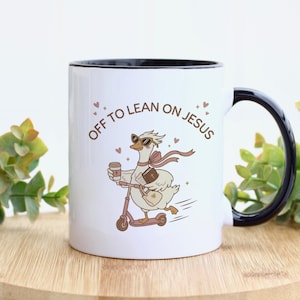 May include: A white ceramic mug with a black rim and handle featuring a cartoon illustration of a goose wearing sunglasses, riding a scooter, holding a coffee cup and a book, with the brown text "OFF TO LEAN ON JESUS" and small hearts.