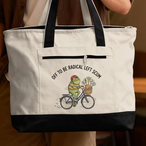 May include: A canvas tote bag with a black base and handles. The bag features a cartoon frog riding a bicycle with a basket full of items. Text on the bag reads "OFF TO BE RADICAL LEFT SCUM."
