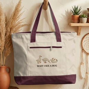 May include: A canvas tote bag with a cream-colored body, a dark burgundy bottom, and matching handles. The front pocket features a zipper and an illustration of three cartoon geese with the text "WAIT I SEE A BUG".