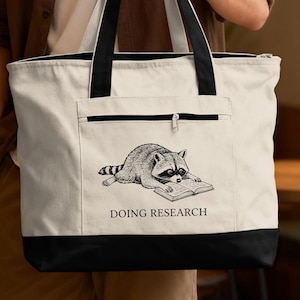 May include: White tote bag with black accents, featuring a raccoon illustration reading a book. The raccoon is in black and white, and the text "DOING RESEARCH" is below the illustration. The bag has a zippered pocket and black handles.