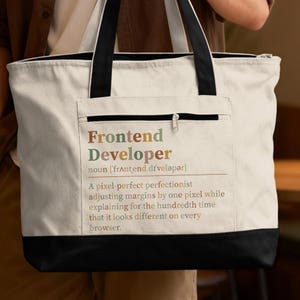May include: A cream-colored canvas tote bag with black handles and a black base. The front features a zippered pocket with the printed text: Frontend Developer noun [frʌntɛnd dɪˈvɛləpər] A pixel-perfect perfectionist adjusting margins by one pixel.