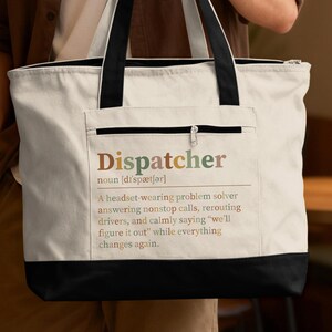 May include: A cream-colored canvas tote bag with black handles and a black base. The front features a zippered pocket printed with the text: Dispatcher noun [dr'spætʃər] A headset-wearing problem solver answering nonstop calls, rerouting drivers.