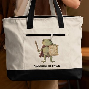 May include: White tote bag with black accents featuring a cartoon frog wearing a cape and holding a staff and a map. The text "We guide at dawn" is printed at the bottom of the bag.