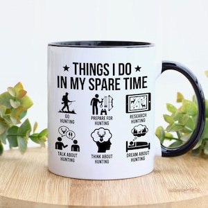 May include: White ceramic mug with a black interior and handle. The mug features the text "THINGS I DO IN MY SPARE TIME" with illustrations depicting hunting activities such as going hunting, preparing, researching, talking, thinking, and dreaming about hunting.