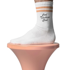 Könnte beinhalten: Weiße Crew-Socken mit zwei orangefarbenen Streifen am oberen Rand und dem Text "Cool Teachers Club" in schwarzer Schrift. Die Socken werden an einem Fuß getragen, der auf einem pfirsichfarbenen Podest ruht.