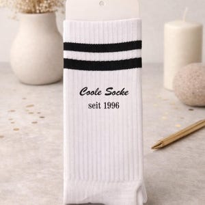 Könnte beinhalten: Weiße gerippte Socken mit zwei schwarzen Streifen am oberen Rand. Der Text "Coole Socke seit 1996" ist schwarz aufgedruckt. Die Socken werden auf einer weißen Karte präsentiert.