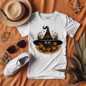 May include: White t-shirt featuring a Halloween-themed graphic of a witch's hat with glowing eyes, surrounded by orange flowers and decorative swirls. The shirt is displayed with a straw hat, sunglasses, and white sneakers on an orange surface.