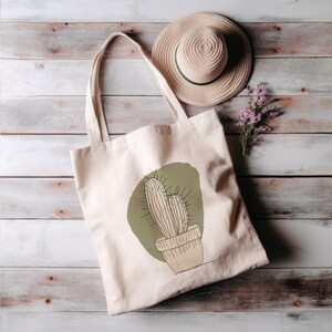 May include: A beige tote bag featuring a green and brown cactus illustration. The bag is on a wooden surface with a straw hat and purple flowers.