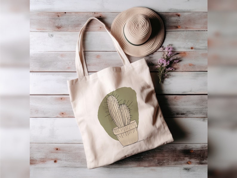 May include: A beige tote bag featuring a green and brown cactus illustration. The bag is on a wooden surface with a straw hat and purple flowers.