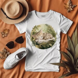 Puede incluir: Camiseta blanca con un gr&aacute;fico de un gato durmiendo en un entorno verde y frondoso. El texto "Fantasy World" est&aacute; impreso en la camiseta.