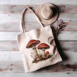 May include: A white canvas tote bag with a design of three red and white mushrooms on a green background. The bag has two long straps and is lying on a wooden surface.