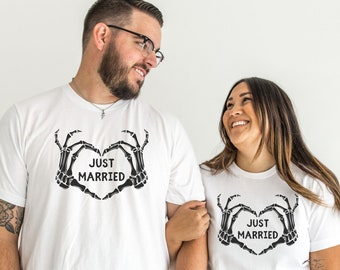 Może przedstawiać: Białe koszulki z czarnym wzorem serca z rąk szkieletu i napisem "JUST MARRIED". Koszulki są noszone przez parę, co sugeruje motyw ślubu lub rocznicy. Projekt jest wyśrodkowany na klatce piersiowej.