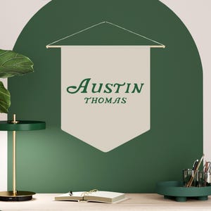 May include: A white banner with the text "Austin Thomas" in green cursive lettering hangs on a green wall. The banner is attached to a wooden dowel rod. A desk with a wooden top and a green chair is in the foreground.