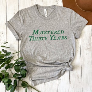 May include: Gray t-shirt with the text "Mastered Thirty Years" in green lettering.