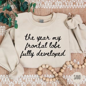 May include: A sand-colored sweatshirt with the text "the year my frontal lobe fully developed" in black script. The sweatshirt is a crew neck style and is made of a soft material. The word "SAND" is in a circle in the bottom right corner.